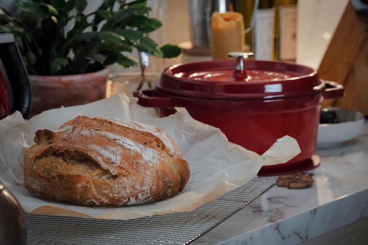 A scored round sourdough loaf on parchment beside a red enamelled cast-iron Dutch oven