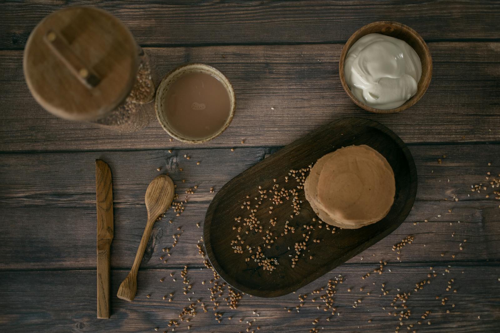 A top-down view of golden-brown buckwheat pancakes on a wooden table with scattered buckwheat grains and a small bowl of sour cream.