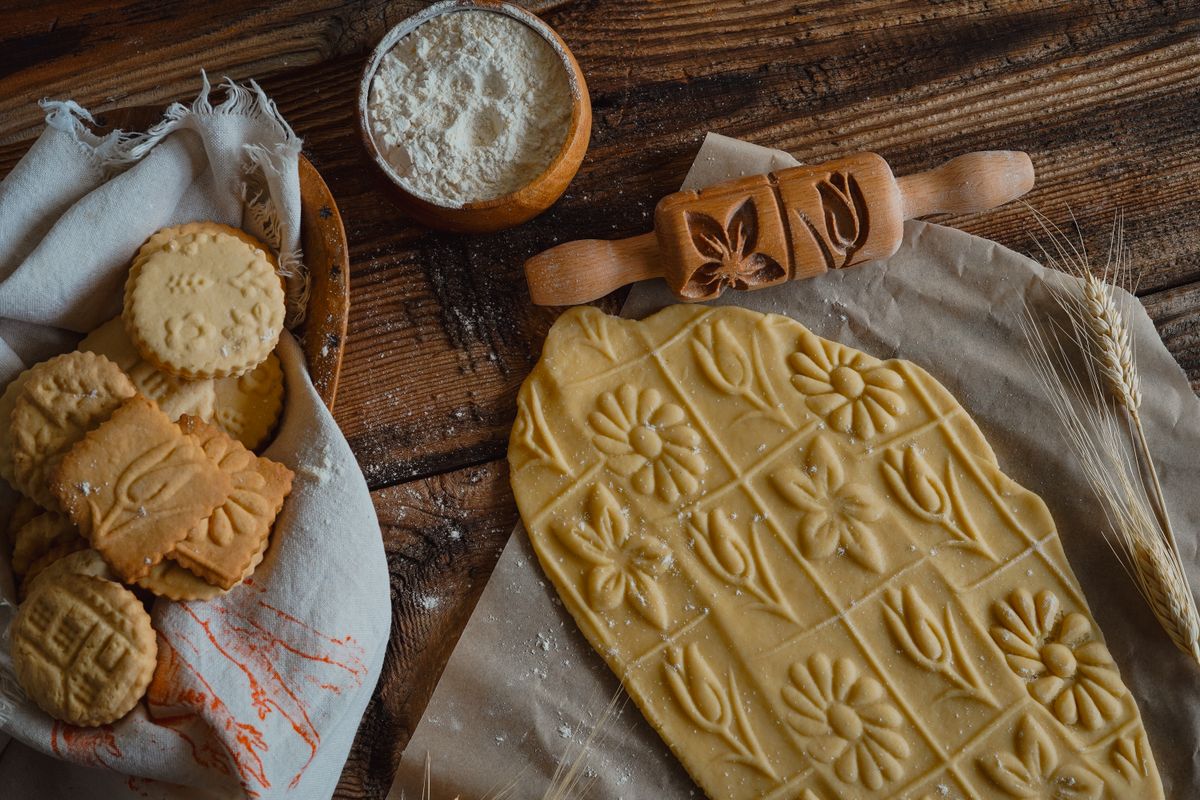 Rolled cookie dough with a patterned rolling pin, baked biscuits in a cloth, a bowl of flour and wheat stalks on a wooden table