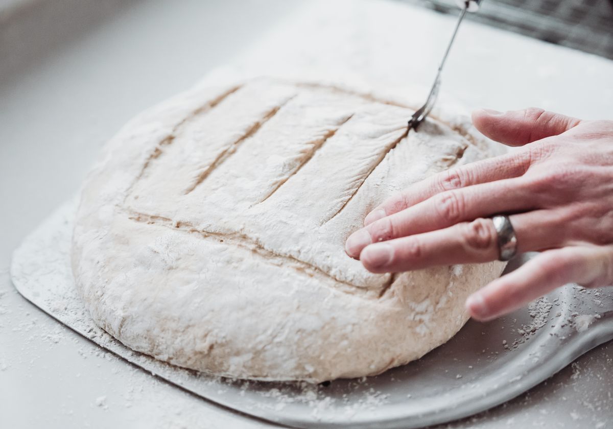 A hand scoring a floured sourdough boule before it goes into the oven.