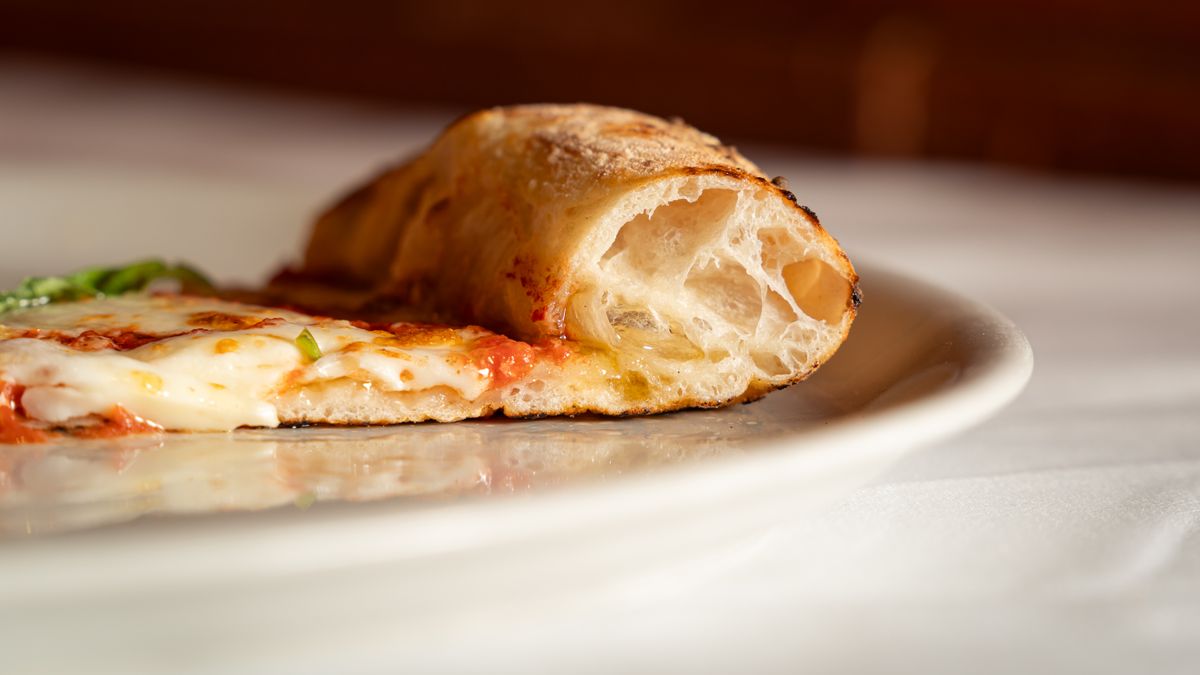 A slice of thin-crust pizza on a white plate showing the airy, charred crust in cross-section.