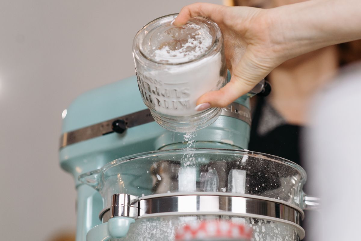 A hand pouring flour from a glass jar, the everyday act of feeding a sourdough starter.