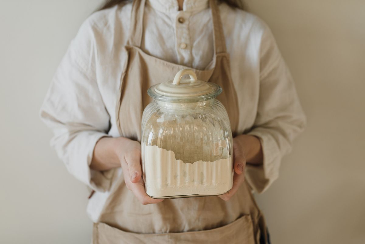 Hands holding a glass jar of flour in a home kitchen, the same kind of jar a new sourdough starter lives in for its first week.