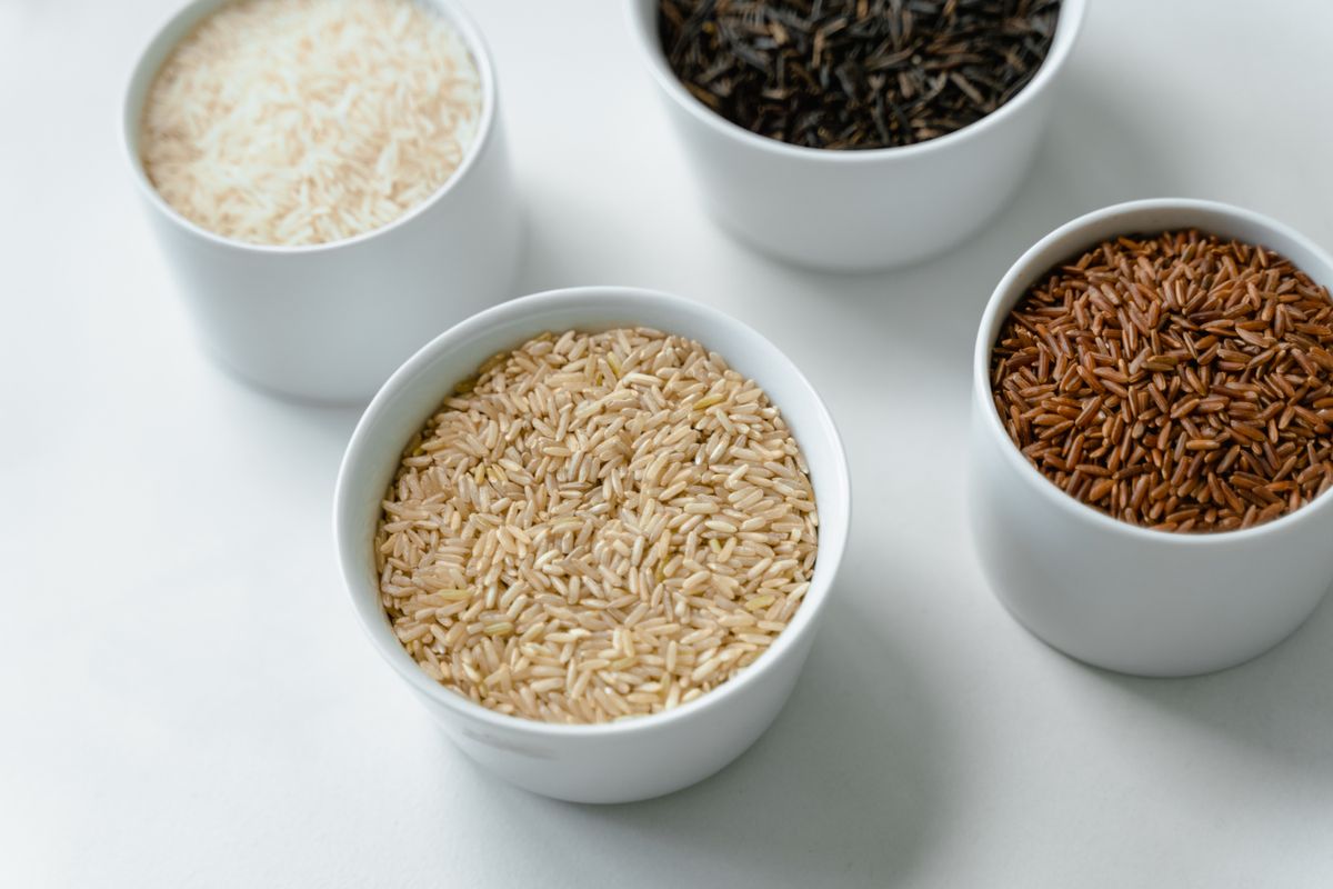 Four white ceramic bowls holding white, brown, wild black, and red rice varieties on a bright surface.