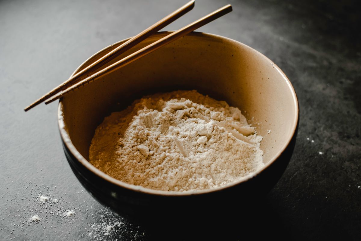 A ceramic bowl of pale rice flour with chopsticks resting across the rim on a dark slate surface.