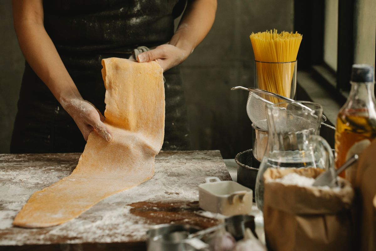 A cook stretches a long golden sheet of fresh pasta dough over a floured wooden board.
