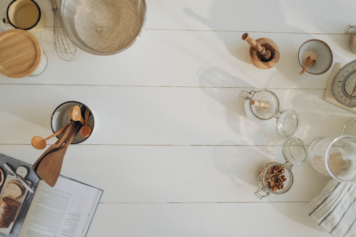 A flat lay of sourdough baking tools, glass jars, flour, and a bread book on a white countertop.